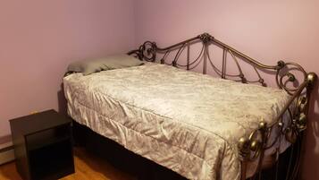 Una cama individual con estructura metálica y una colcha estampada en una habitación con piso de madera y una mesita de noche negra pequeña.