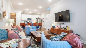 Ein modernes Wohnzimmer mit Sofa, Couchtisch und Fernseher. Im Hintergrund ist eine Küche mit weißen Schränken und ein Essbereich mit orangefarbenen Stühlen zu sehen.