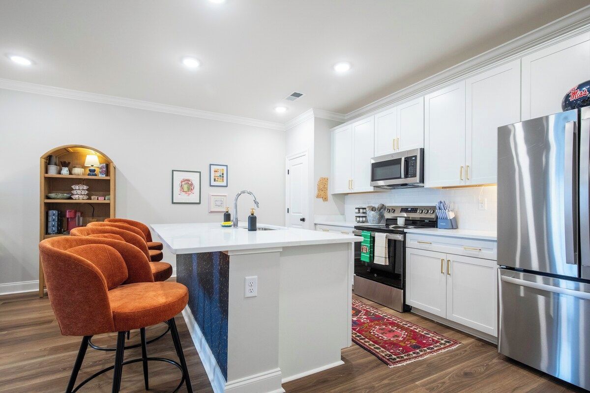A modern kitchen with white cabinets, a stainless steel refrigerator, and a bar area with orange chairs.