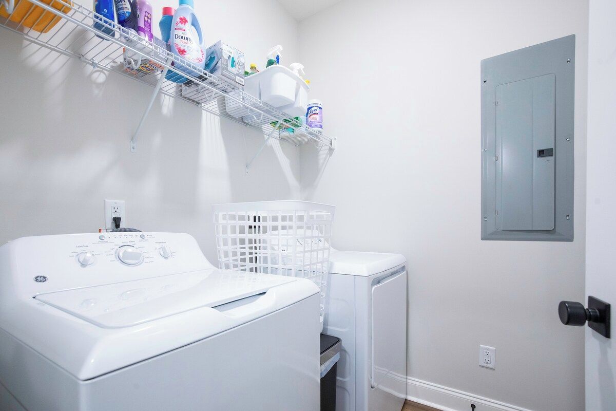 A laundry area with a front-loading washer and dryer, a white laundry basket, and a shelf with various cleaning products.