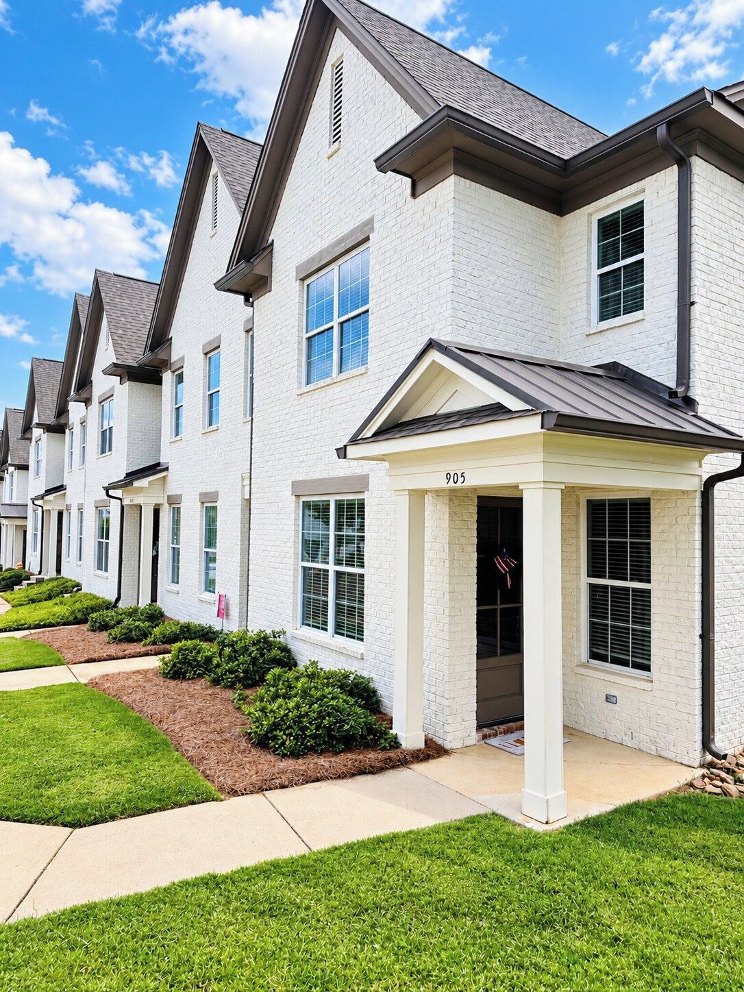 A row of modern townhouses with a uniform architectural style, featuring white brick exteriors, dark roofs, and a central entrance with a covered porch.