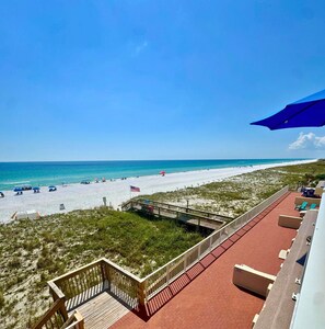Blick auf einen Strand von einem Balkon mit Holzgeländer, roten Fliesenböden und einem wolkenlosen blauen Himmel.