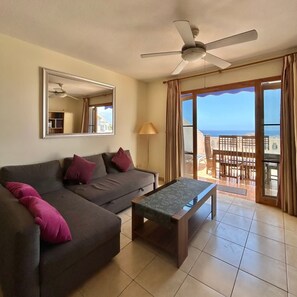 Ein Wohnzimmer mit Sofa, Couchtisch, Deckenventilator, einem großen Fenster mit Vorhängen und Blick auf den Ozean.