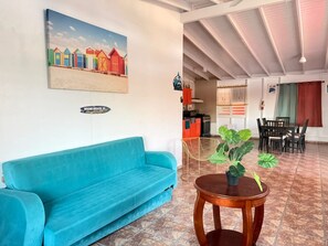 A room with a blue sofa, a wooden table with a potted plant, a painting of beach huts, and a kitchen area in the background. - Spacious & Colorful Haven with Pool Tables & Pool (Cabo Rojo)