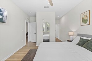 A bedroom with a large bed, a bedside table with a lamp, a door leading to another room, and a window with a view of greenery. - Largo 2BR Hideaway | Free Parking, Pool & Gym (Largo)