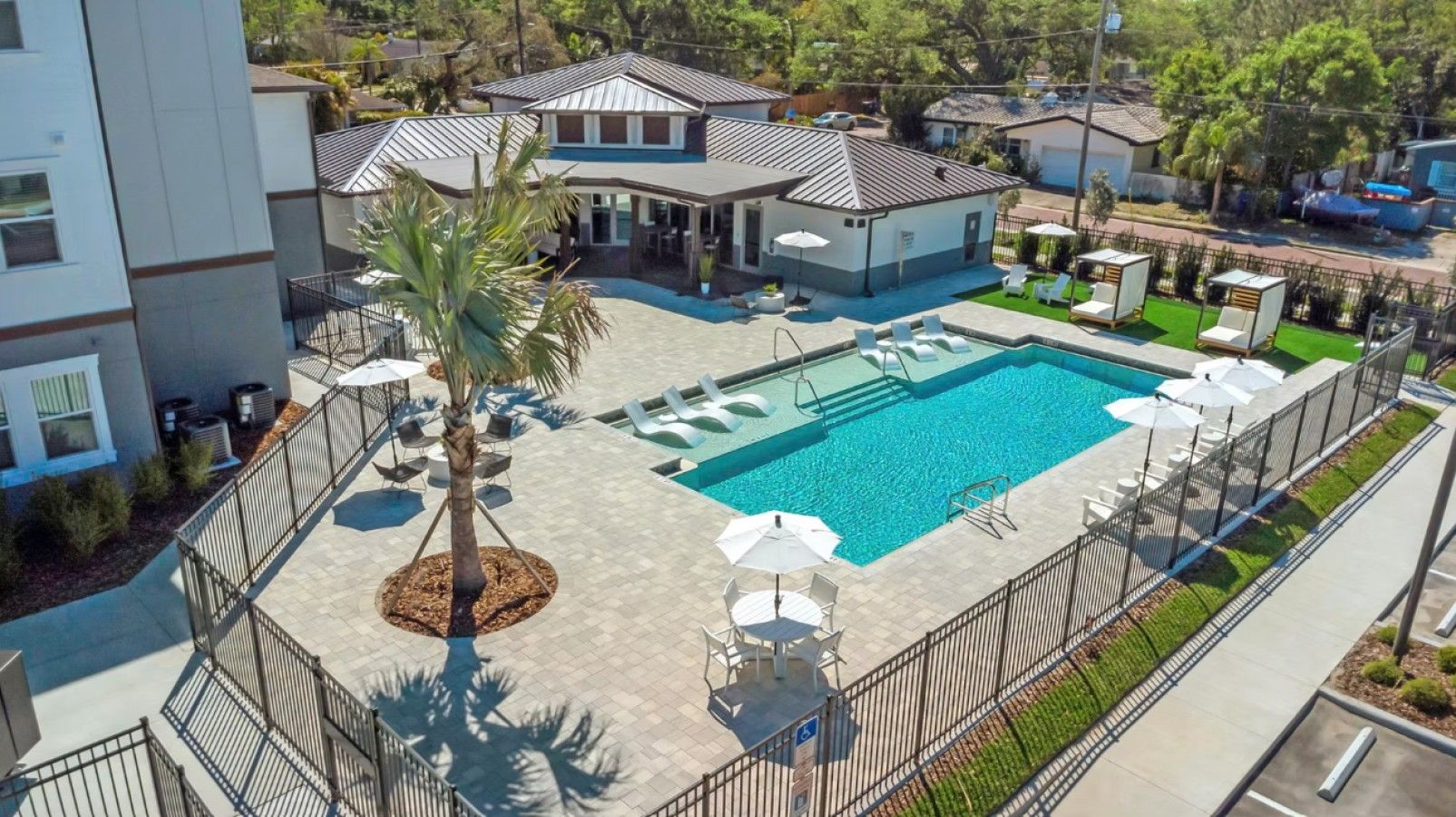 A swimming pool with lounge chairs and umbrellas, surrounded by a fence and palm trees.