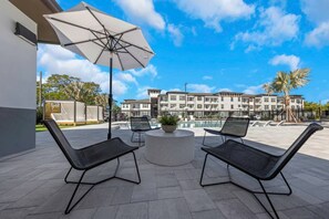 An outdoor seating area with two chairs and a table, a large umbrella, and a building with multiple balconies in the background. - Relaxing 2BR Stay | Pool, Gym & Free Parking (Largo)
