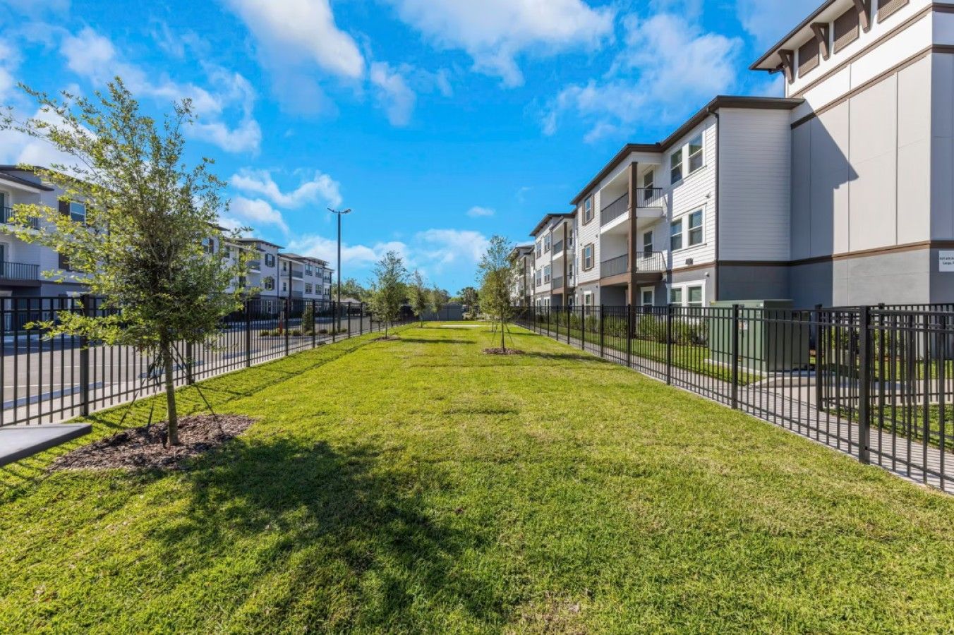 A residential area with a well-maintained lawn, a black metal fence, and multi-story apartment buildings.