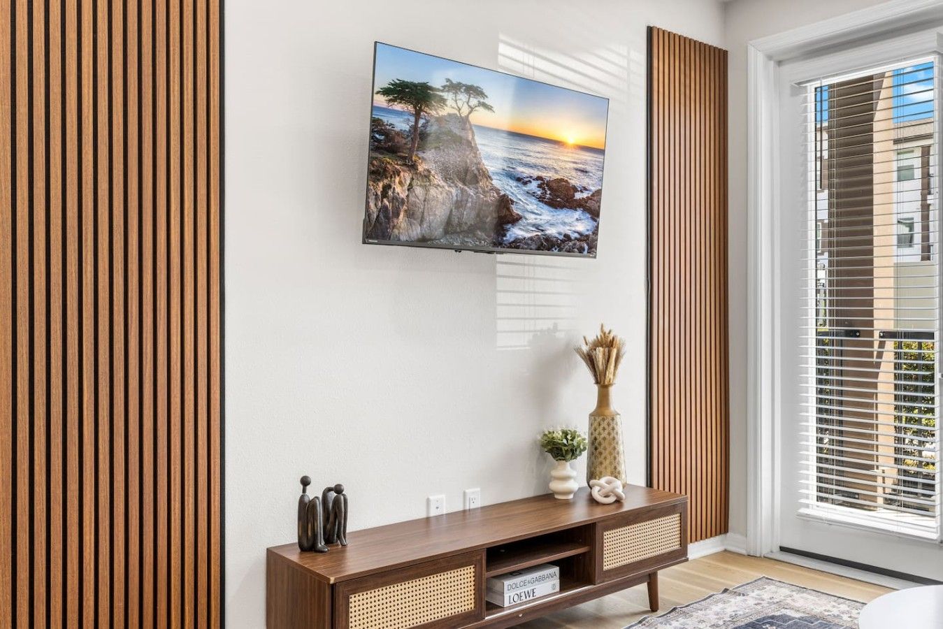 Un salon avec un téléviseur fixé au mur, des lambris en bois, une console ornée d’objets décoratifs et une vue par la fenêtre.