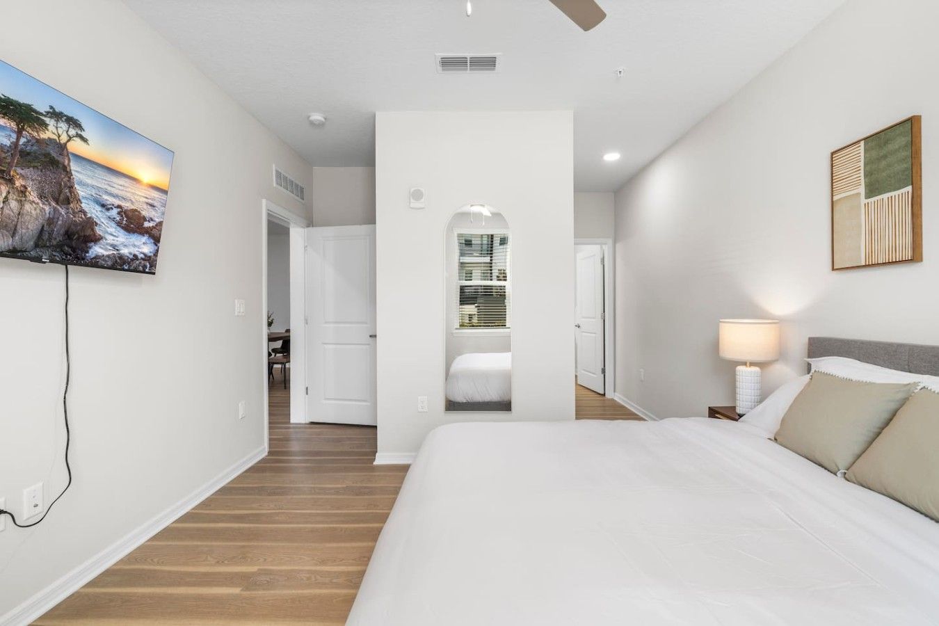 A bedroom with a large bed, a nightstand, a lamp, and a painting of a coastal scene.
