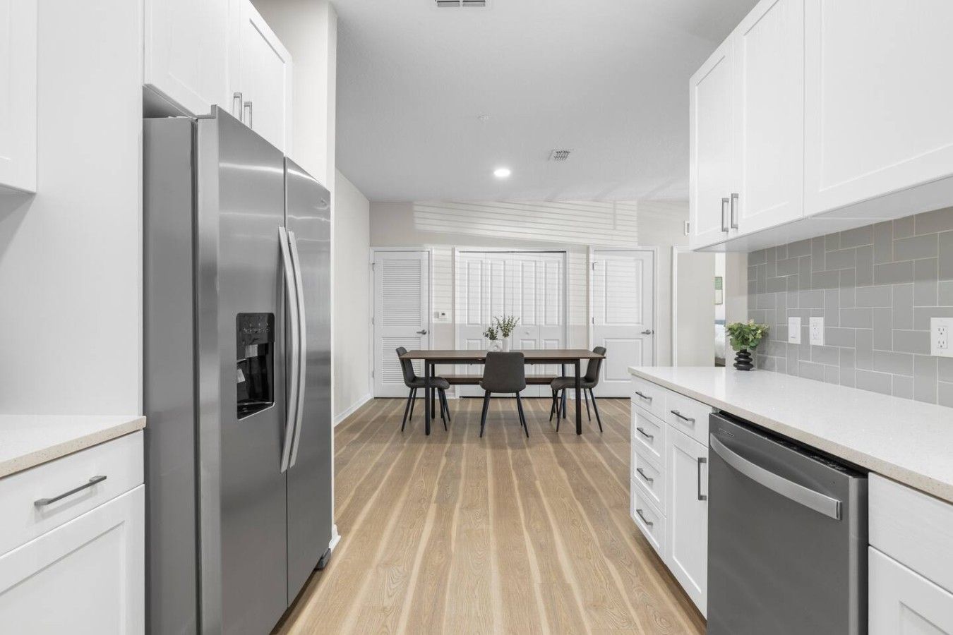 A modern kitchen with white cabinetry, a stainless steel refrigerator, a dining table with chairs, and a dishwasher.