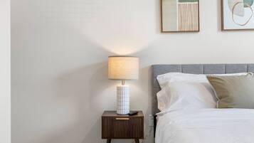 Una cama bien hecha con ropa blanca y un cabecero gris, una mesita de noche con lámpara, y dos cuadros enmarcados en la pared.