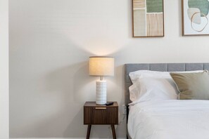 A neatly made bed with white bedding and a grey headboard, a bedside table with a lamp, and two framed artworks on the wall. - Relaxing 2BR Stay | Pool, Gym & Free Parking (Largo)