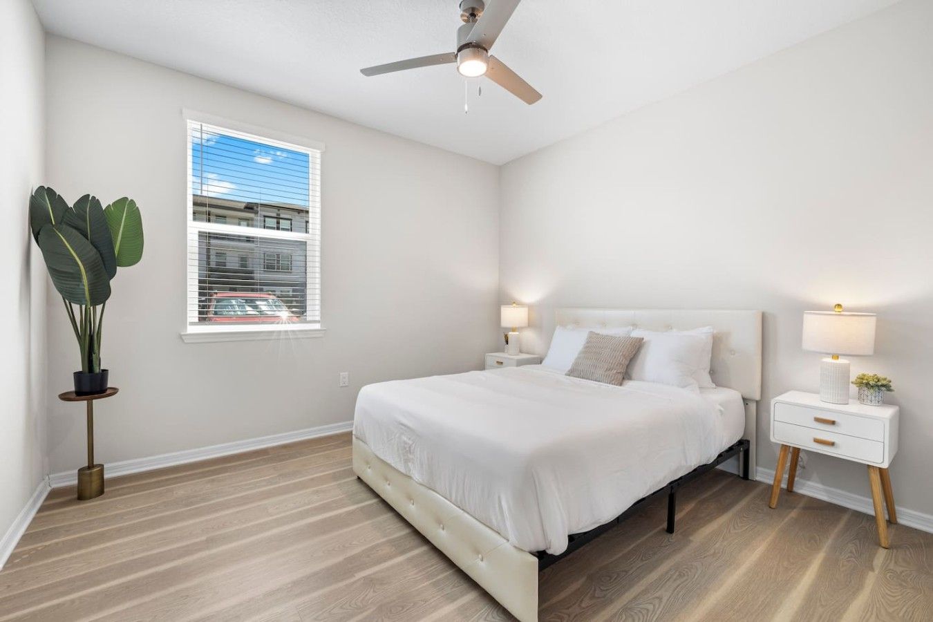 A bedroom with a bed, bedside tables, a ceiling fan, a window with blinds, and a potted plant.