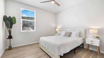 A bedroom with a bed, bedside tables, a ceiling fan, a window with blinds, and a potted plant.