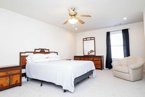 Ein Schlafzimmer mit einem Bett, einem Schminktisch, einem Sessel, einem Deckenventilator und einem Fenster mit Vorhängen.