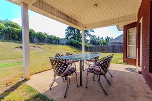 Eine überdachte Terrasse mit einem Glastisch und Stühlen, im Hintergrund ein Ziegelhaus und ein eingezäunter Garten.