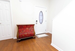 Quarto com piso de madeira, cômoda vermelha com estampas florais, porta branca com painel de vidro e parede branca.