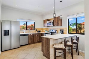 A modern kitchen with a central island, stainless steel appliances, wooden cabinets, and a view through large windows. - Villa Sail Away | Private Jacuzzi & Courtyard (Noord)