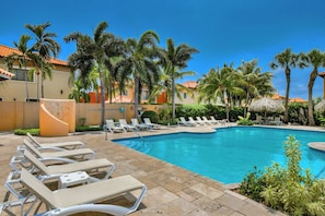 A swimming pool with lounge chairs, palm trees, and a building with a tiled roof. - Villa Sail Away | Private Jacuzzi & Courtyard (Noord)