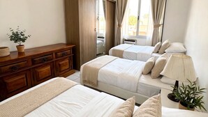 Une chambre d’hôtel avec deux lits, une commode en bois, une fenêtre avec des rideaux, une lampe de chevet et une plante en pot.