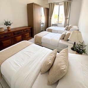 Habitación de hotel con dos camas, un aparador de madera, una ventana con cortinas, una lámpara de noche y una planta en maceta.