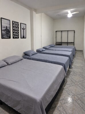 Quarto de hotel com quatro camas dispostas em linha, um ventilador de teto e três quadros na parede.