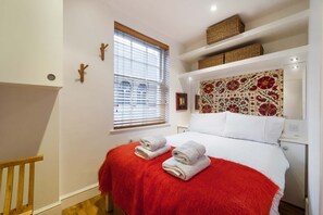 A bedroom with a bed, a window with blinds, a red throw blanket, and a decorative headboard. - Cosy 1-bed - Fitzrovia - Sleeps 3 (London)
