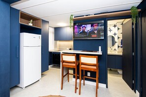 A compact kitchen with a refrigerator, two wooden chairs, a small island, and a TV screen displaying a movie. - SHB - Studio Grego com Pé na Areia (Santos)