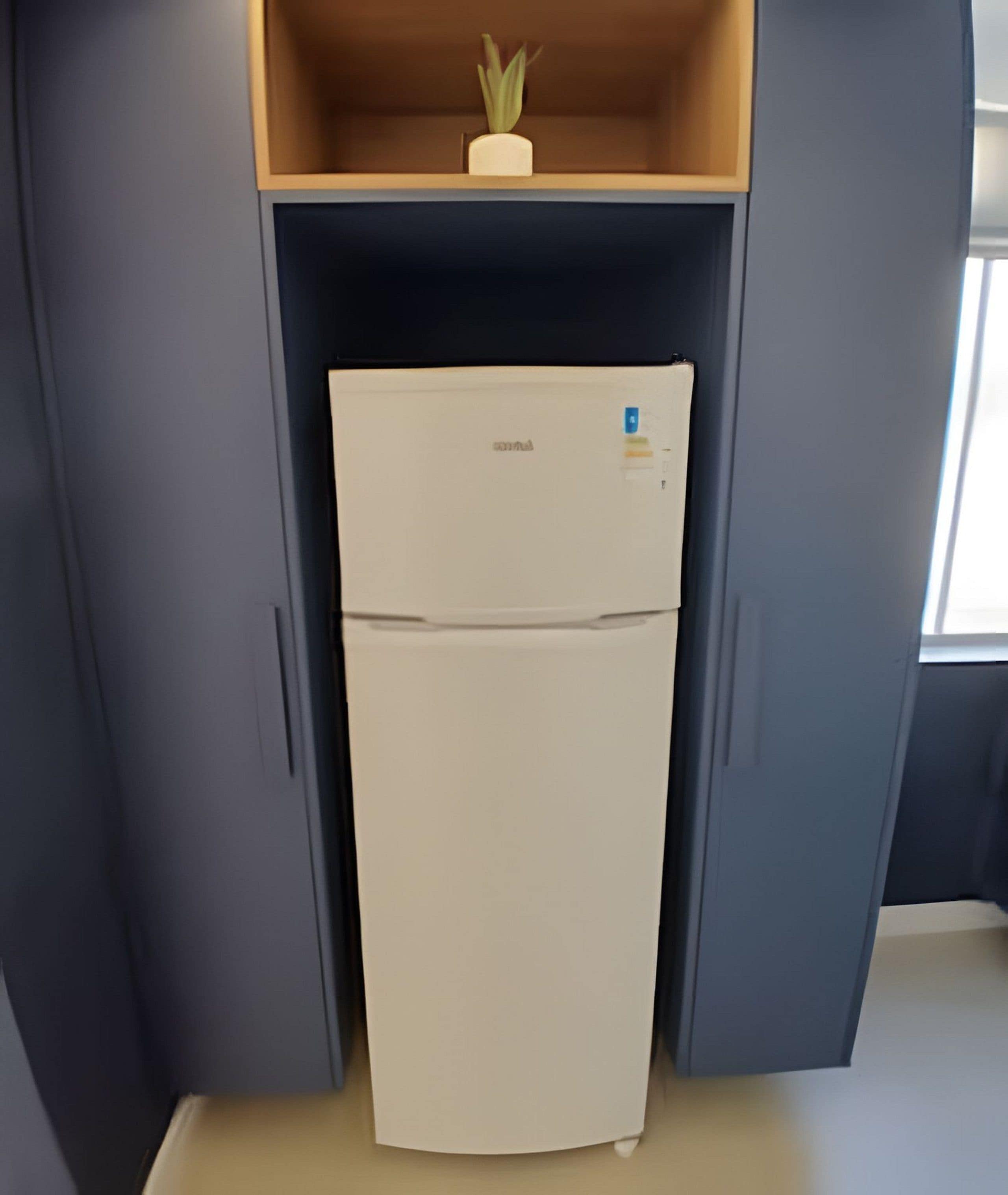 Un refrigerador compacto con una pequeña planta encima, ubicado en un mueble estrecho con paredes grises.