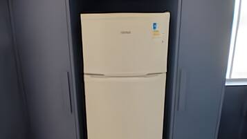 Un refrigerador compacto con una pequeña planta encima, ubicado en un mueble estrecho con paredes grises.