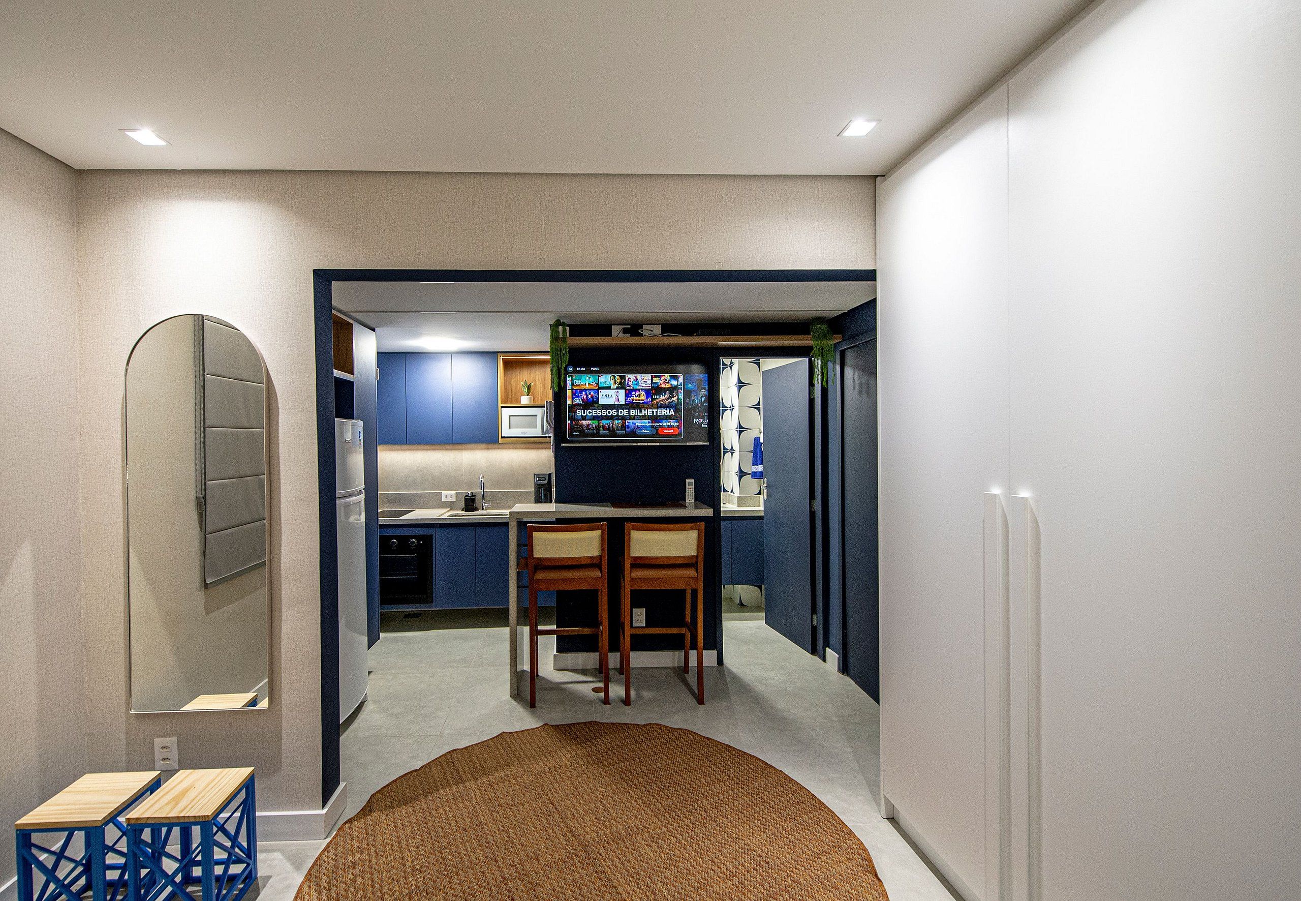 Een moderne keuken met blauwe kastjes, een eethoek met twee houten stoelen en een grote flatscreen televisie bevestigd boven de aanrecht.