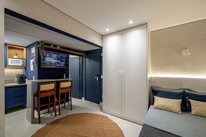 A compact bedroom with a bed, a small desk with a chair, a microwave, and a television. - SHB - Studio Grego com Pé na Areia (Santos)