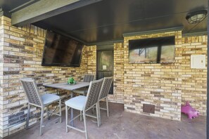 Eine Terrasse im Freien mit einer Ziegelwand, einem Tisch mit Stühlen und zwei Wandmonitoren.