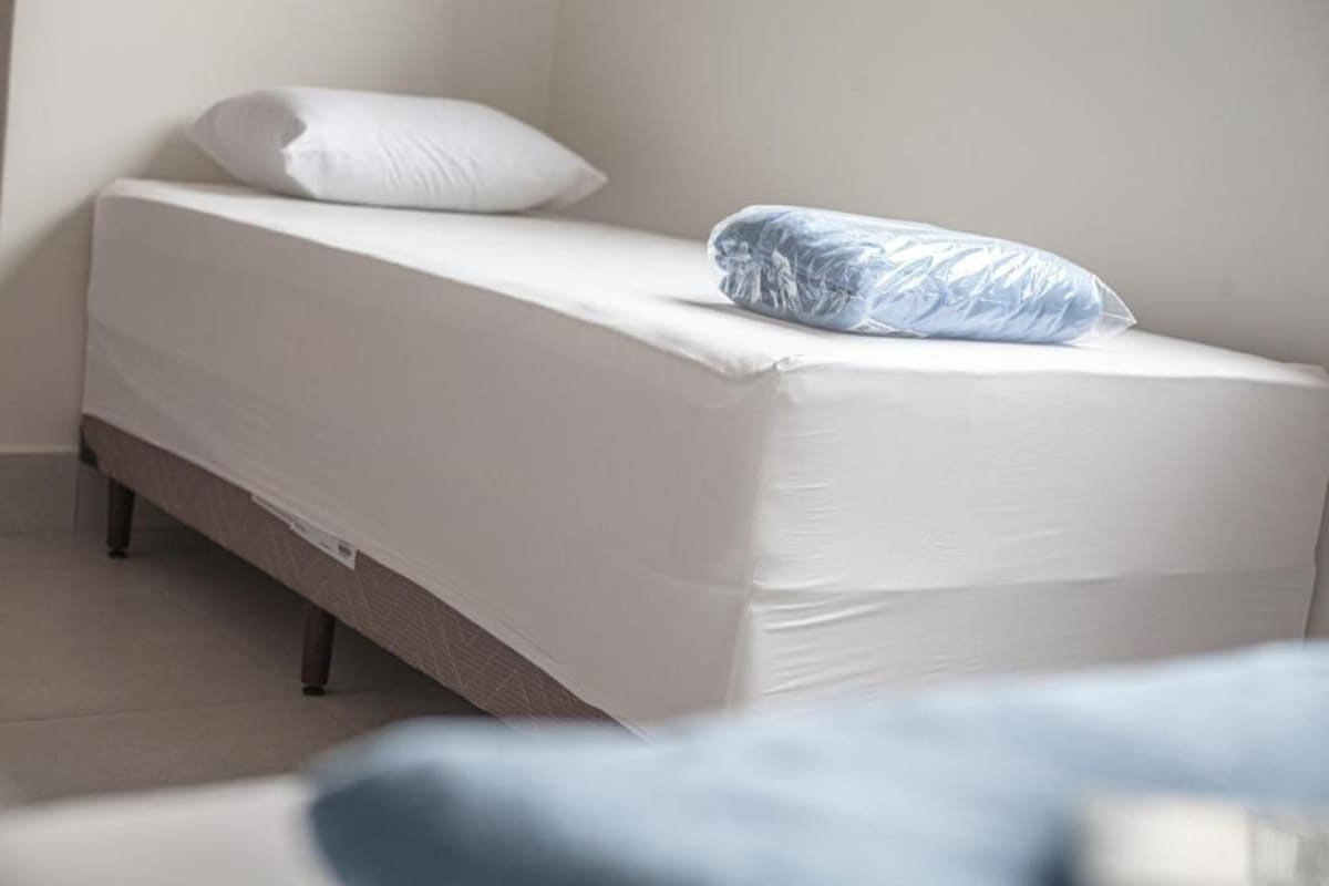 Una cama individual con colchón y almohada blancos, cabecera de madera y pared blanca.