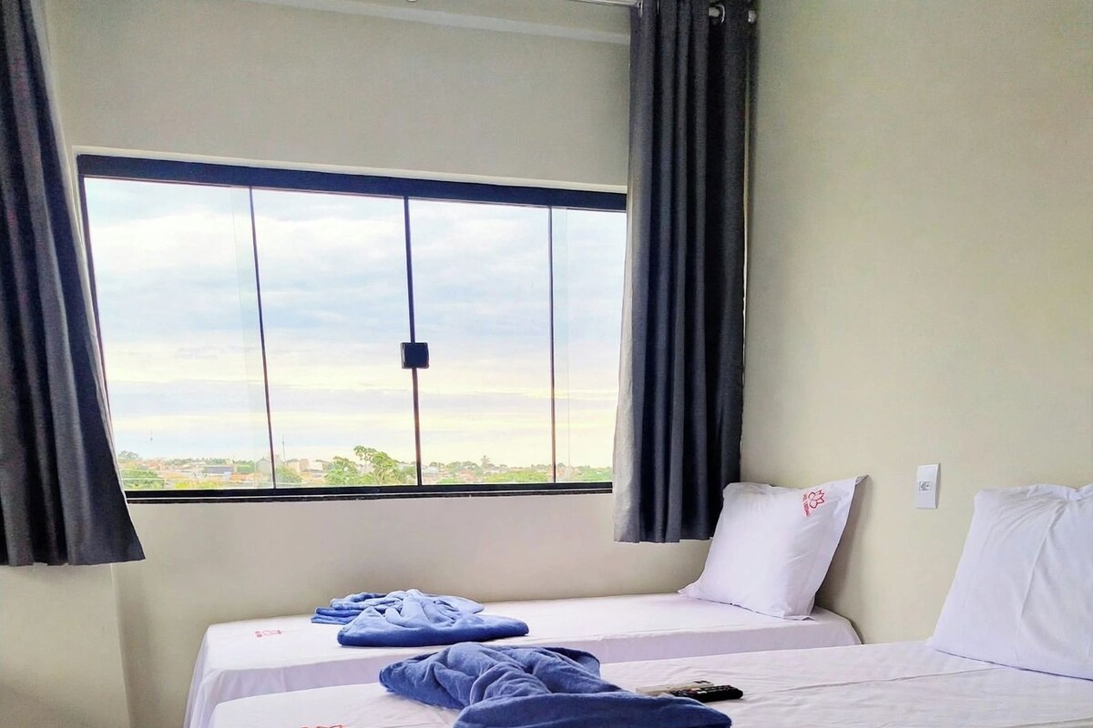 Una habitación de hotel con dos camas, cada una con una colcha blanca y una toalla azul. Hay una ventana con cortinas y el suelo es de baldosas.
