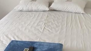 Una cama con ropa de cama blanca y dos almohadas, una manta azul con un control remoto y una pared lisa.