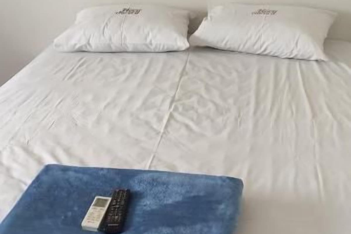 Una cama con ropa de cama blanca y dos almohadas, una manta azul con un control remoto y una pared lisa.