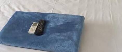 A bed with white bedding and two pillows, a blue blanket with a remote control, and a plain wall.