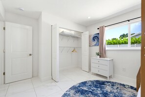 A bedroom with a large window, a wardrobe, a bedside table, and a blue patterned rug. - Our Caribbean Home (West Palm Beach)