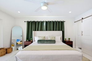 A bedroom with a bed, a ceiling fan, a large mirror, and a view of the beach. - Our Caribbean Home (West Palm Beach)