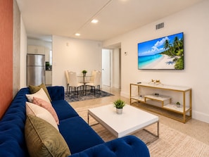 A modern living room with a blue sofa, a white coffee table, and a painting of a beach scene. - 5-Star Location | Heart of Wynwood 2BR+Parking (Miami)