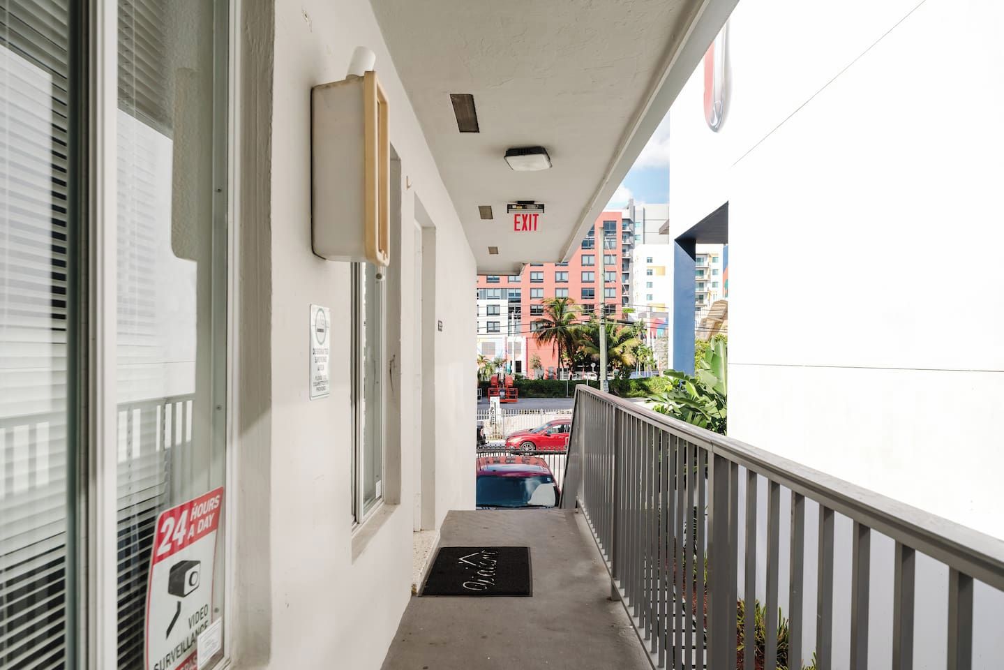 Un couloir étroit avec une rambarde en métal, une porte portant un panneau d’avertissement de sécurité, et une vue sur un parking avec des voitures et des bâtiments en arrière-plan.