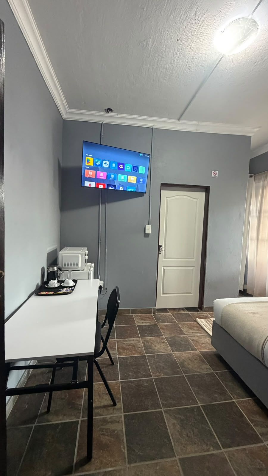 Een kleine kamer met een bed, een bureau met stoel, een aan de muur bevestigde televisie en een deur.