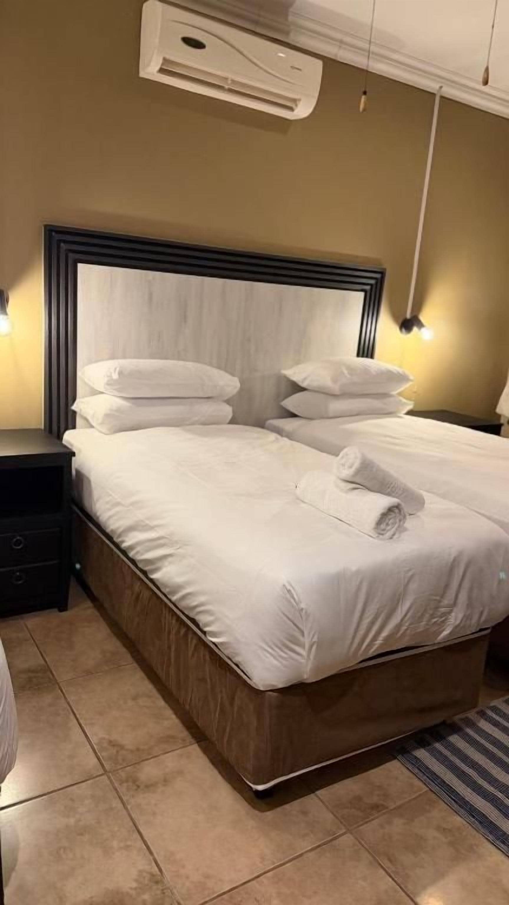 Una cama bien hecha con ropa blanca y una toalla doblada, una mesita de noche y un equipo de aire acondicionado instalado en la pared.