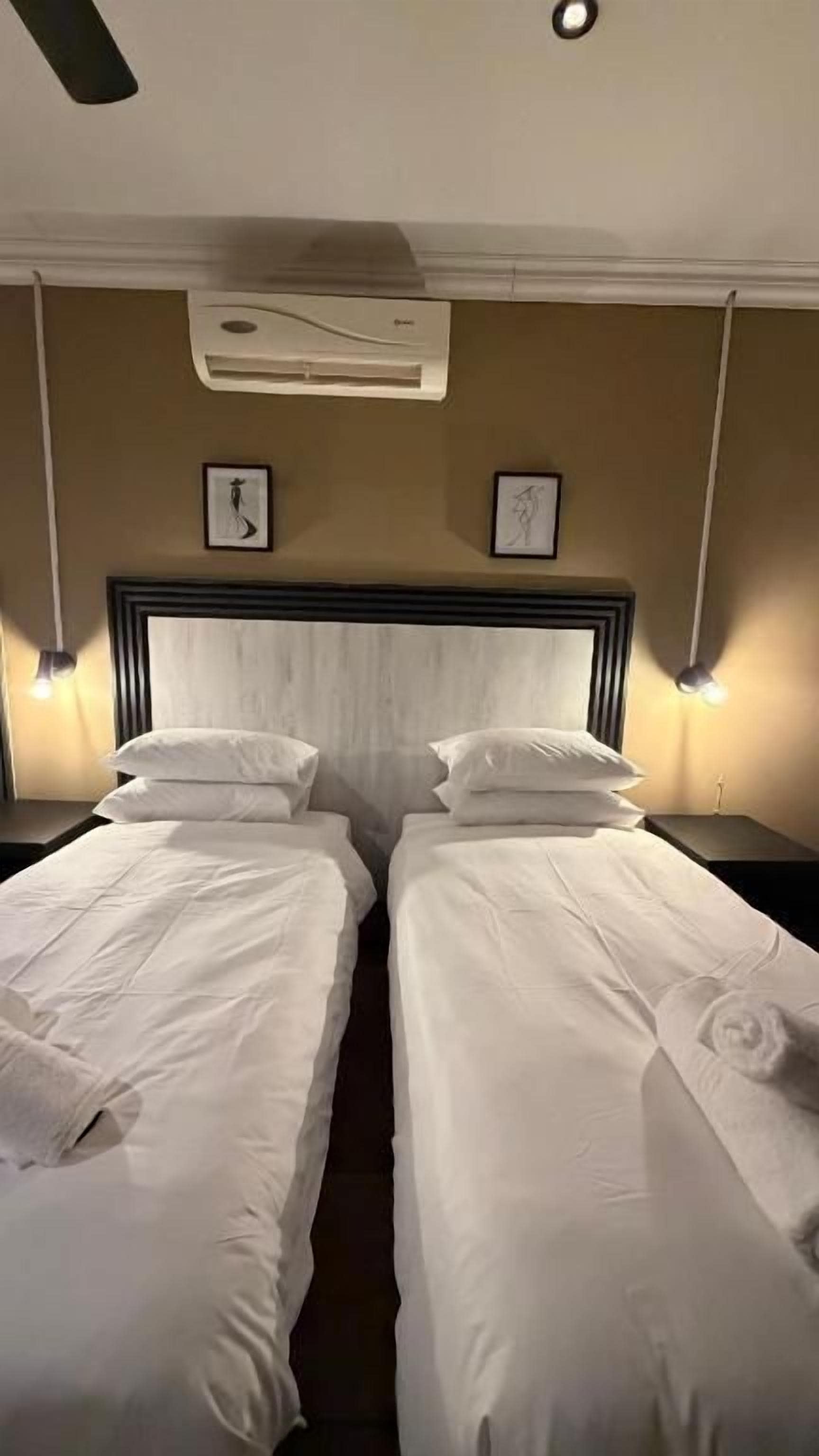 A hotel room with two beds, a headboard, wall-mounted air conditioning, and two wall-mounted lamps.