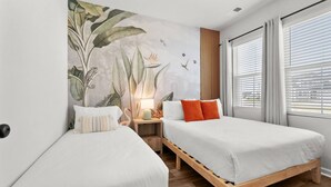 Et hotelværelse med to senge, et natbord med lampe, et mural med tropiske planter og et vindue med rullegardiner.