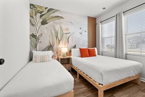 Une chambre d’hôtel avec deux lits, une table de chevet avec une lampe, une fresque représentant des plantes tropicales et une fenêtre avec des stores.