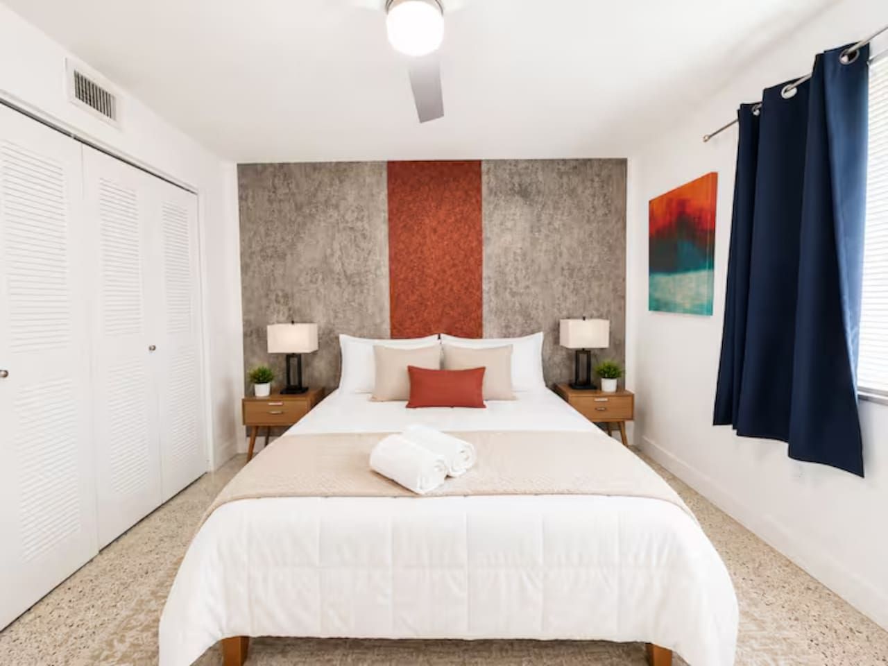 Une chambre d’hôtel avec un grand lit, deux tables de chevet avec des lampes, une tête de lit au motif texturé, une fenêtre avec des rideaux et une porte donnant sur un placard.