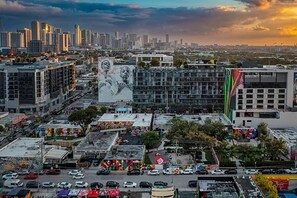 Une vue urbaine avec une grande fresque murale sur un immeuble, entouré de divers bâtiments commerciaux et bordé d’une rue animée.